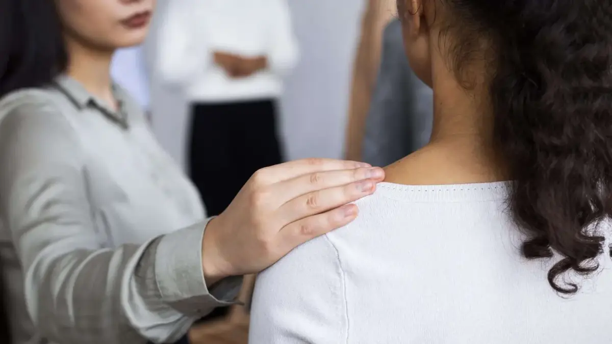Woman consoling another woman during a group therapy session with a hand on the shoulder