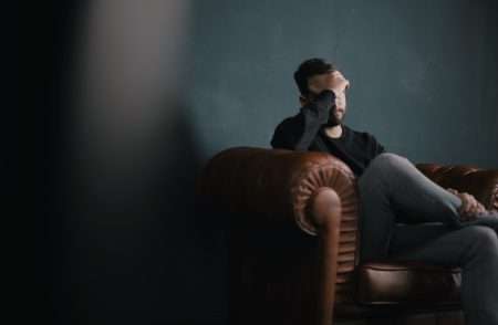 Man sitting in a dark room on a couch with his head resting on his hand