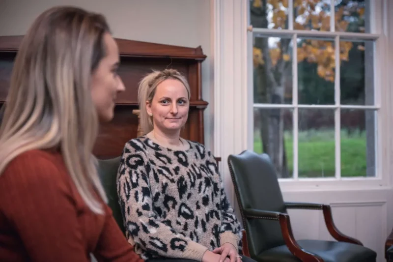 Indoor picture of two women sitting in chairs in a therapy session smiling while offering support to each other