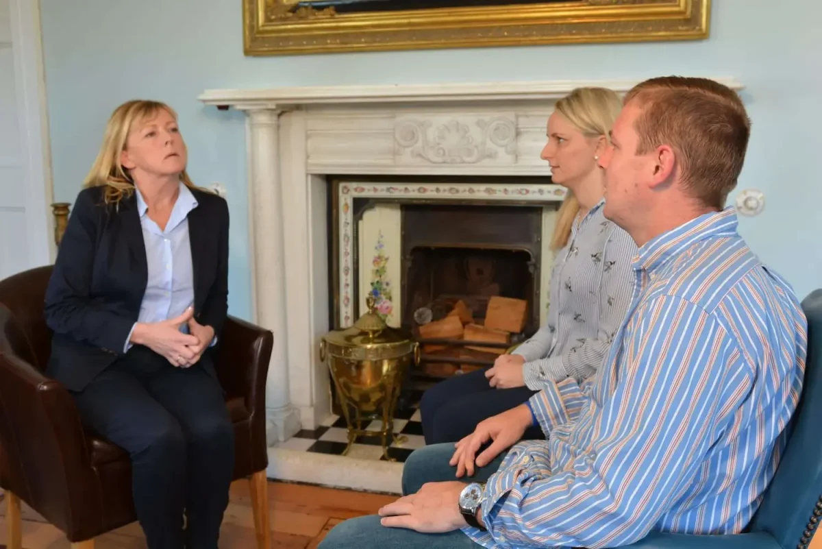 Three people (two women and a man) dressed professionally sat across from one another in conversation indoors in front of a fireplace